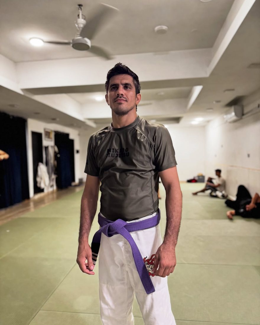 Amjad Khan in a grey rash guard and purple belt, standing on the Mikado mats